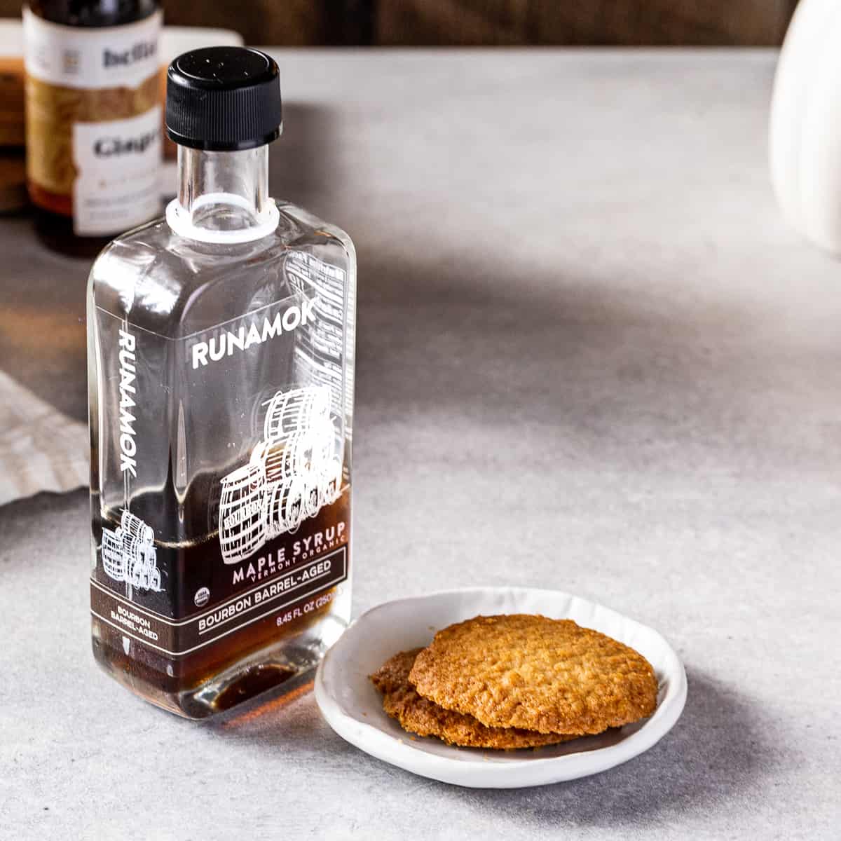 Maple syrup and a dish of cookies together on a countertop.