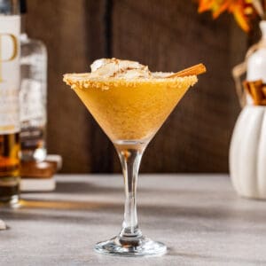 Side view of a Pumpkin Martini cocktail with a whipped cream and cinnamon stick garnish. In the background are fall leaves as well as a white pumpkin holding cinnamon sticks, along with maple syrup and a bottle of bourbon.