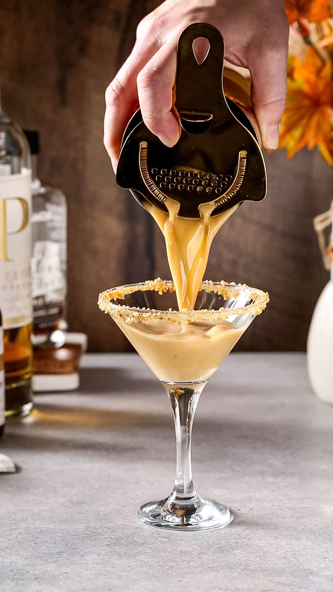 Hand using a cocktail strainer to pour brown liquid into a martini glass that has a cookie crumb coated rim.