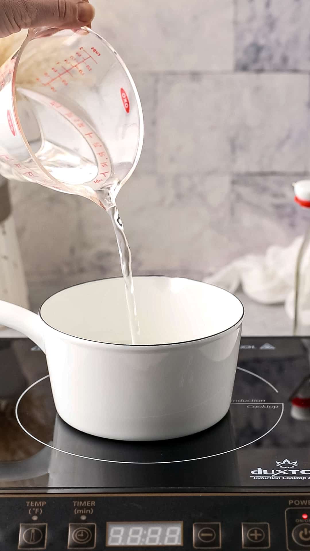 Water being poured from a measuring cup into a white saucepan. The saucepan is on an induction burner.