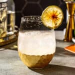 Side view of a Smoky Margarita in a stemless gold-bottomed wine glass. The drink has a salted rim and torched lime garnish. In the background are tequila, smoked syrup and a jigger.