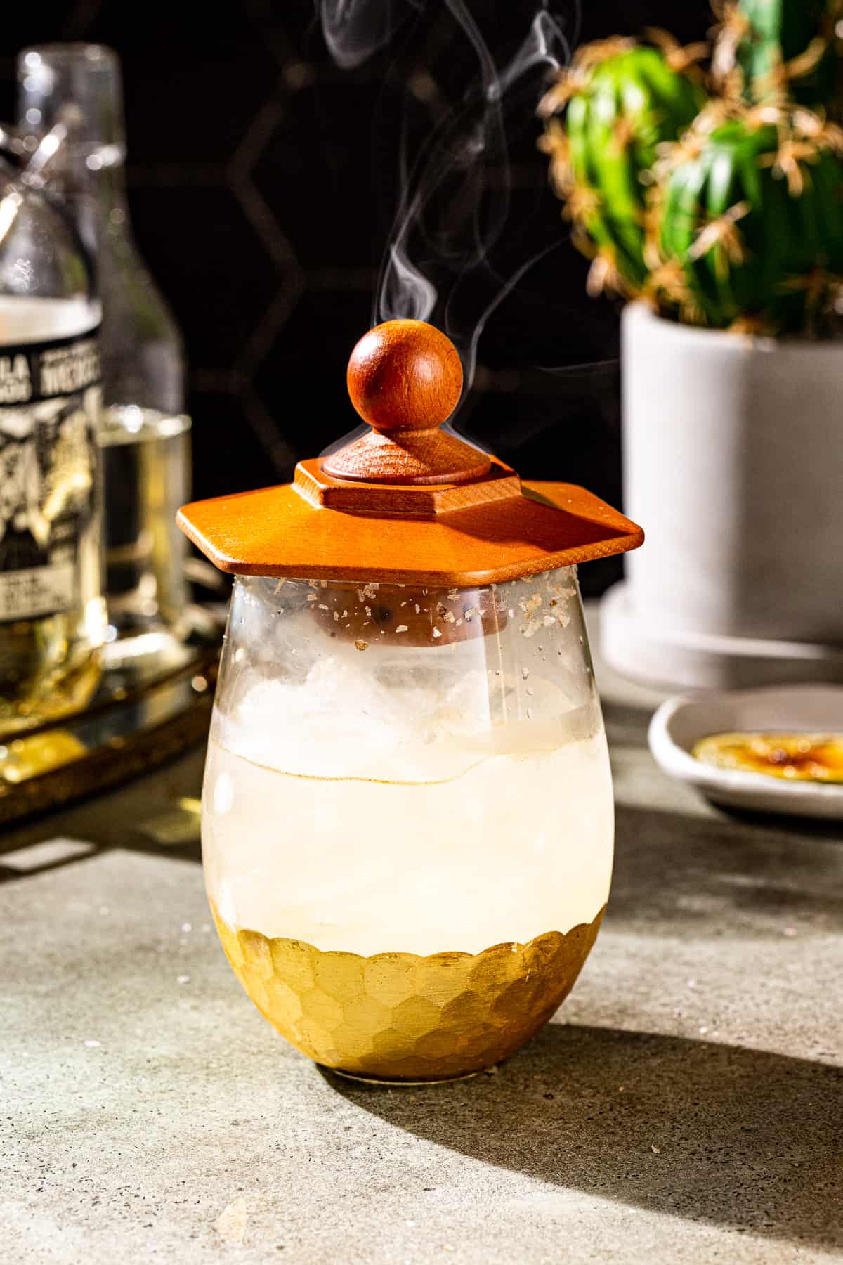 Wooden cocktail smoker on top of the margarita serving glass. The glass is filled with the margarita, ice and smoke, and wisps of smoke are rising from the cocktail smoker.