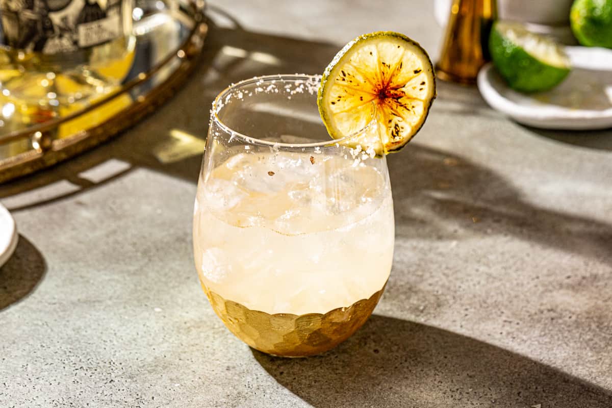 Smoky Margarita cocktail in a gold-bottomed stemless wine glass with a salted rim and a brûléed lime garnish on a countertop. A gold mirrored tray is in the background along with tequila, a jigger and limes.