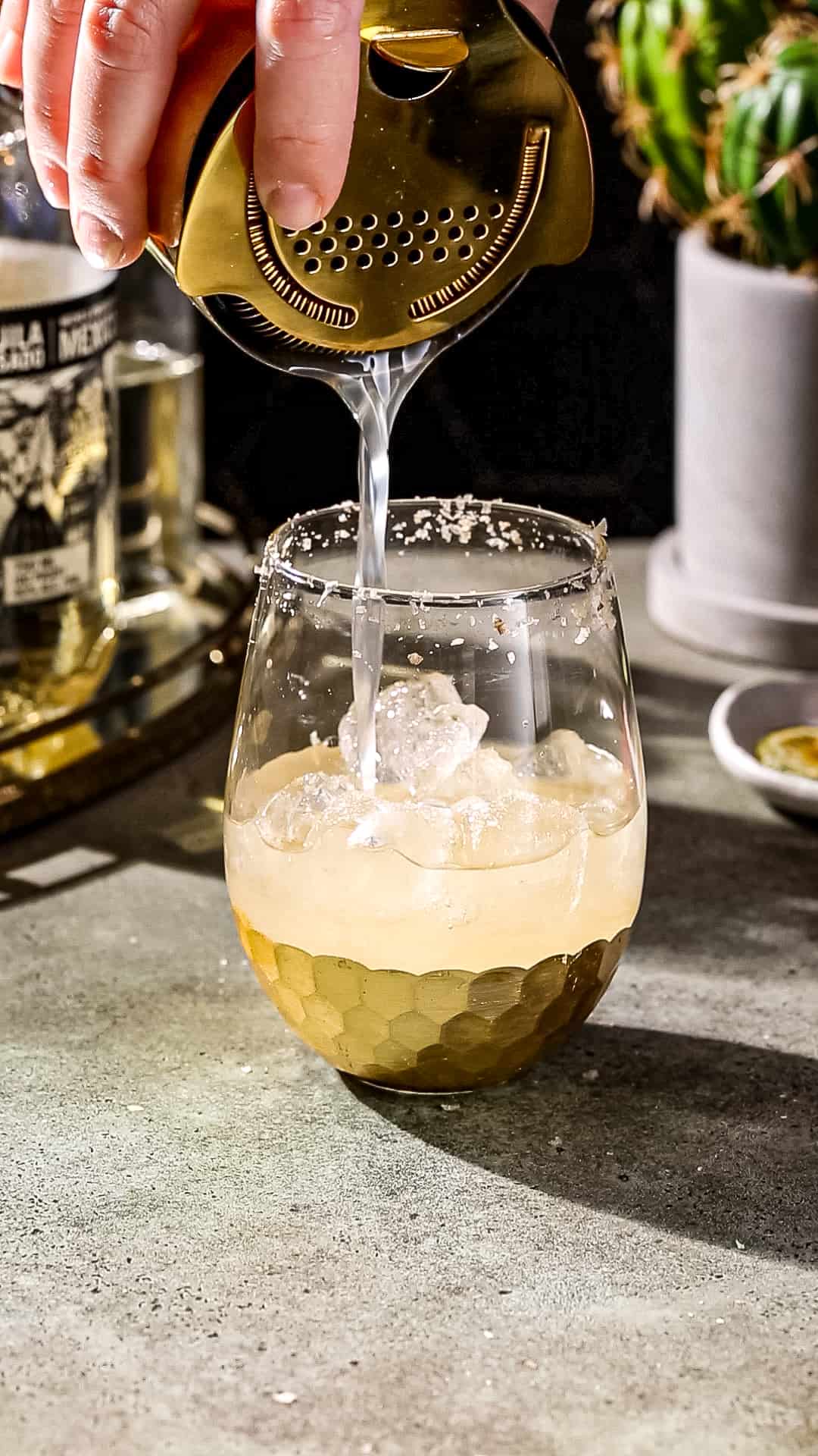 Hand using a gold colored cocktail shaker and strainer to pour a drink into a serving glass that is filled with ice.