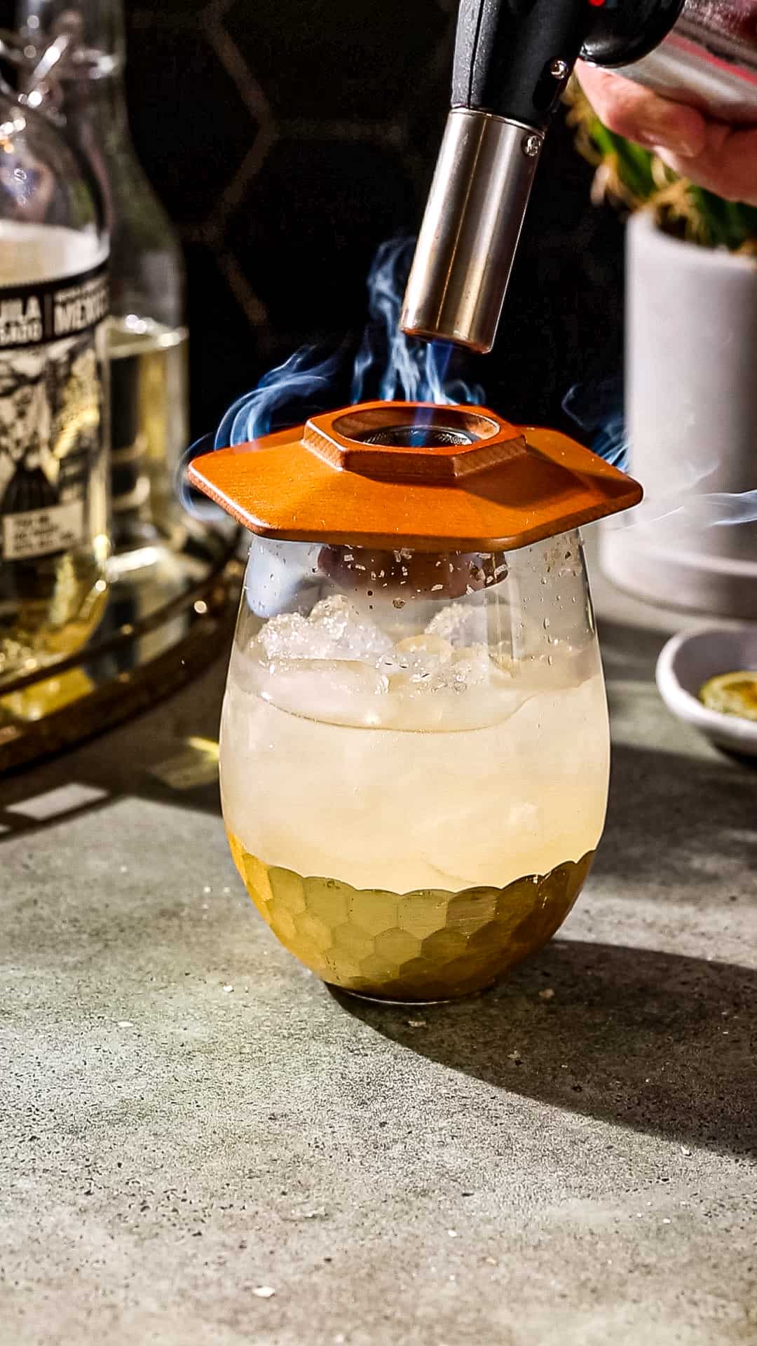 Hand using a kitchen torch to ignite the wood chips in a cocktail smoker that is sitting on top of a cocktail serving glass filled with ice and liquid.