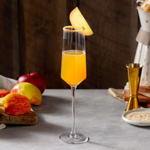 Side view of an Apple Cider mimosa on a gray countertop in a Champagne flute. Cinnamon sugar is coating the rim of the glass and an apple slice is attached to the rim as a garnish. In the background is fresh apple, a jigger, a plate of cinnamon sugar and a plate of caramel sauce, along with a rustic vase.