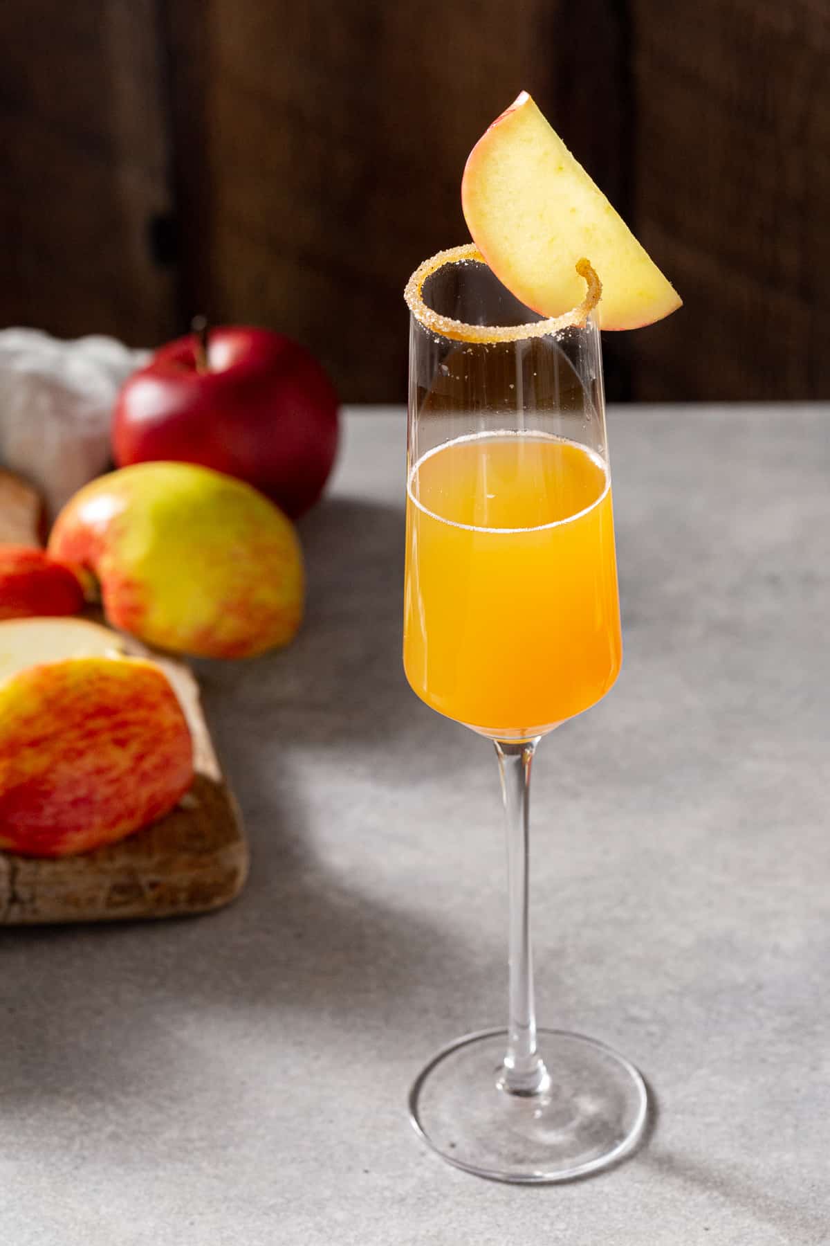 An Apple Cider mimosa on a gray countertop in a Champagne flute. Cinnamon sugar is coating the rim of the glass and an apple slice is attached to the rim as a garnish. In the background to the left of the drink is a pile of cut up fresh apple pieces.