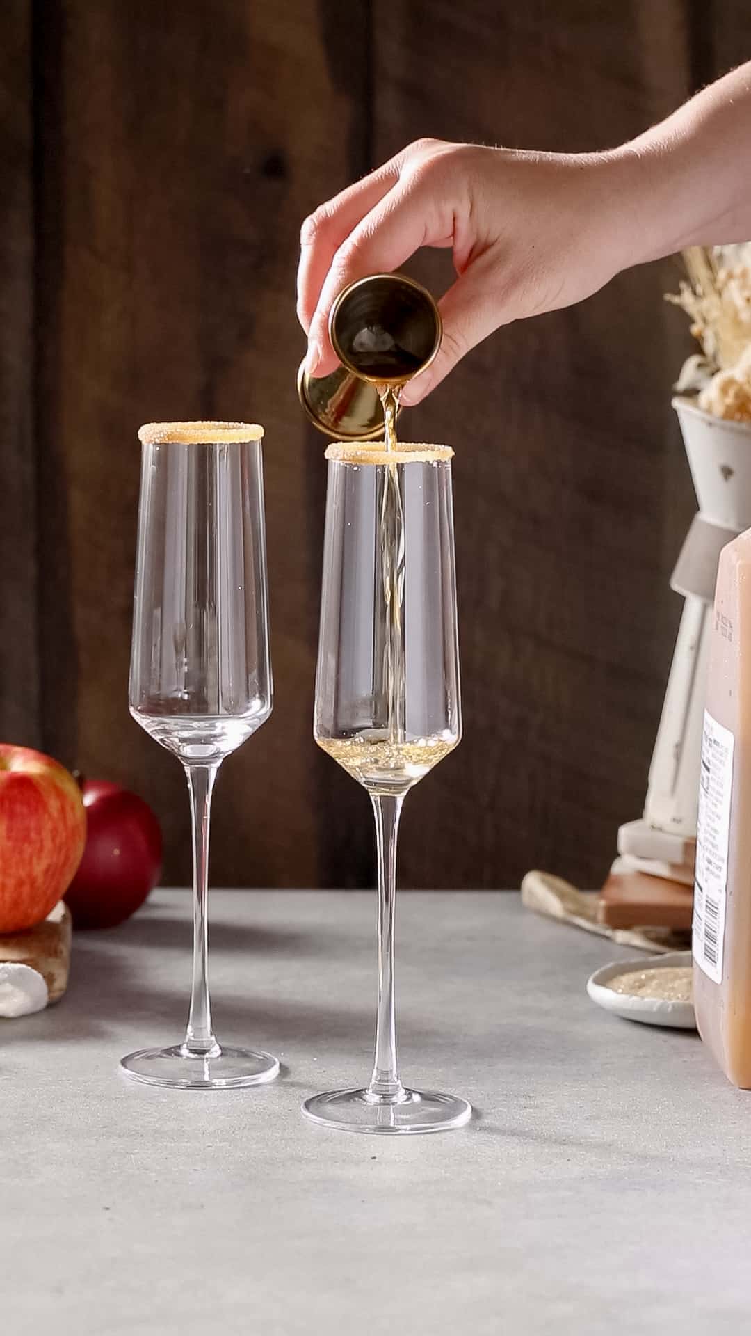 Hand using a gold jigger to pour applejack into a champagne flute. A second champagne flute is slightly in back to the left.