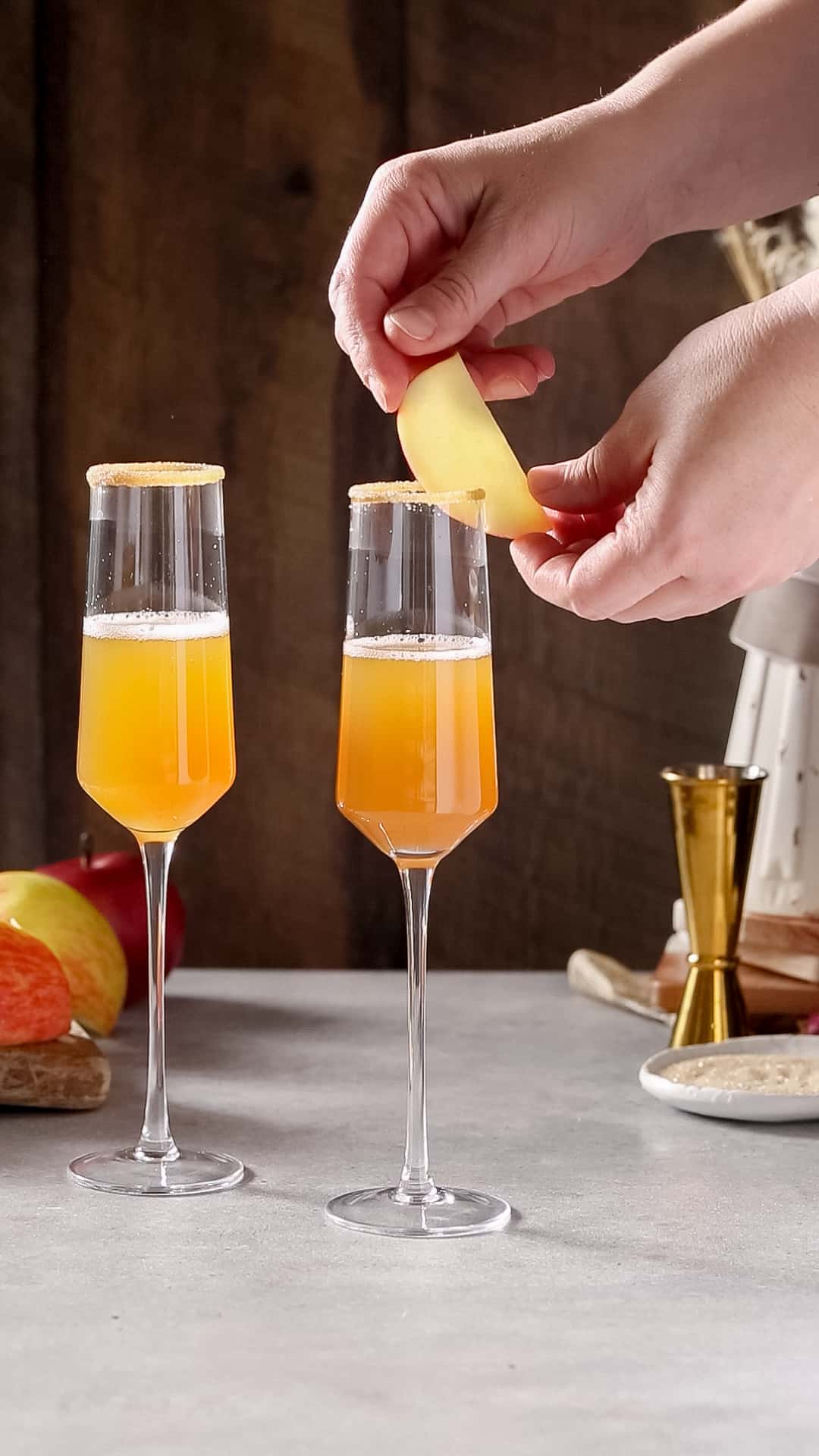 Hand adding an apple slice to the rim of a champagne flute with an Apple Cider Mimosa in it. Another mimosa is in the background to the left.