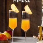 Two Apple Cider mimosas on a gray countertop with a wood grain wall in the background. Cinnamon sugar is coating the rim of the Champagne flutes and an apple slice is attached to each rim as a garnish. In the background is fresh apple, a gold jigger, a plate of cinnamon sugar and a plate of caramel sauce, along with a slightly visible rustic vase. Text above the top of the drinks reads “Apple Cider Mimosa”.