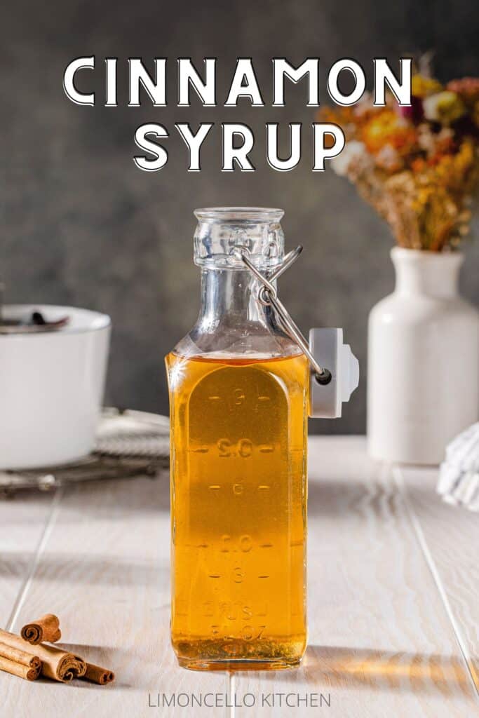 Glass bottle filled with amber-colored cinnamon syrup on a white wood countertop. In the background are a white pot on a cooling rack and a vase with dried flowers. Above the glass bottle is text that reads “Cinnamon Syrup”.