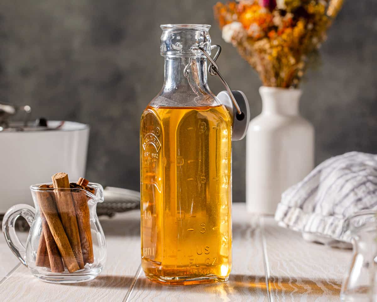 Side view of an open glass bottle filled with cinnamon syrup. A glass container with cinnamon sticks is to the left of the bottle. They are on a white wood countertop and in the background are a white saucepan on a round cooling rack, a white vase with fall-colored dried flowers, and a white and gray striped linen.