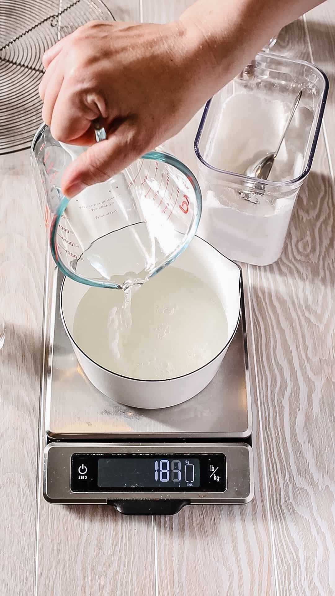 Overhead view of a white saucepan on a kitchen scale. A hand is pouring water from a measuring cup into the saucepan.