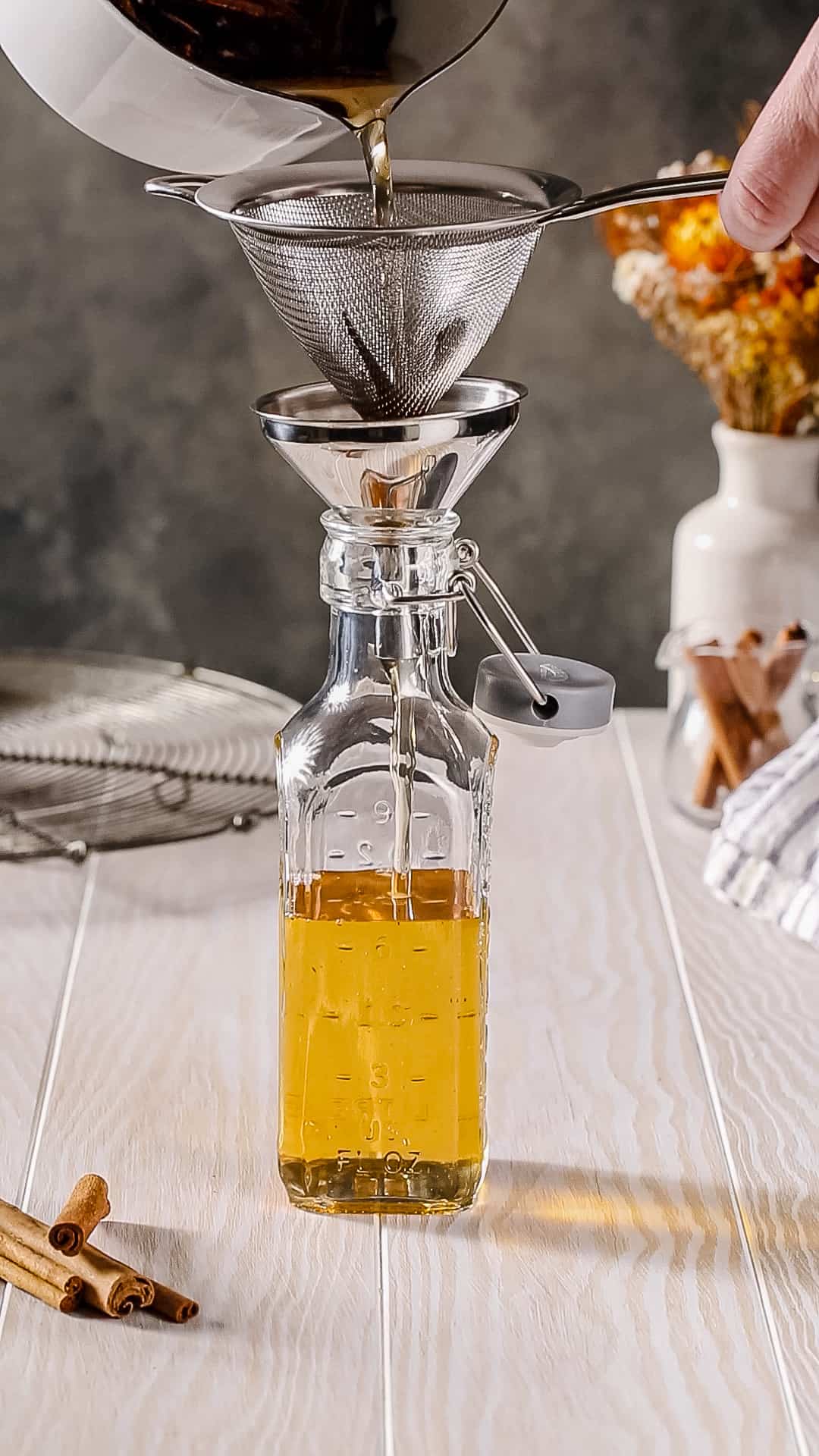 Side view of a glass bottle with syrup being poured into it through a strainer and funnel.