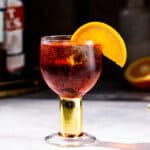 Side view of a Negroni Sbagliato cocktail on a gray countertop. The drink is in a goblet style glass with a short gold stem and an orange slice garnish. The background is in shadow but a cut orange is visible to the right, and a bottle of vermouth and Campari liqueur are slightly visible on the left.