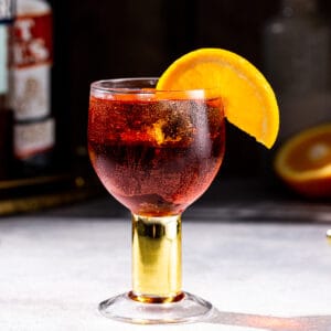 Side view of a Negroni Sbagliato cocktail on a gray countertop. The drink is in a goblet style glass with a short gold stem and an orange slice garnish. The background is in shadow but a cut orange is visible to the right, and a bottle of vermouth and Campari liqueur are slightly visible on the left.