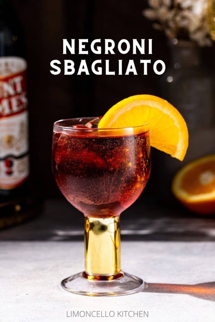 Side view of a Negroni Sbagliato cocktail on a gray countertop. The drink is in a goblet style glass with a short gold stem and an orange slice garnish. The background is in shadow but a cut orange is visible to the right, and a bottle of vermouth is slightly visible on the left. Above the drink, text reads “Negroni Sbagliato” in all caps.