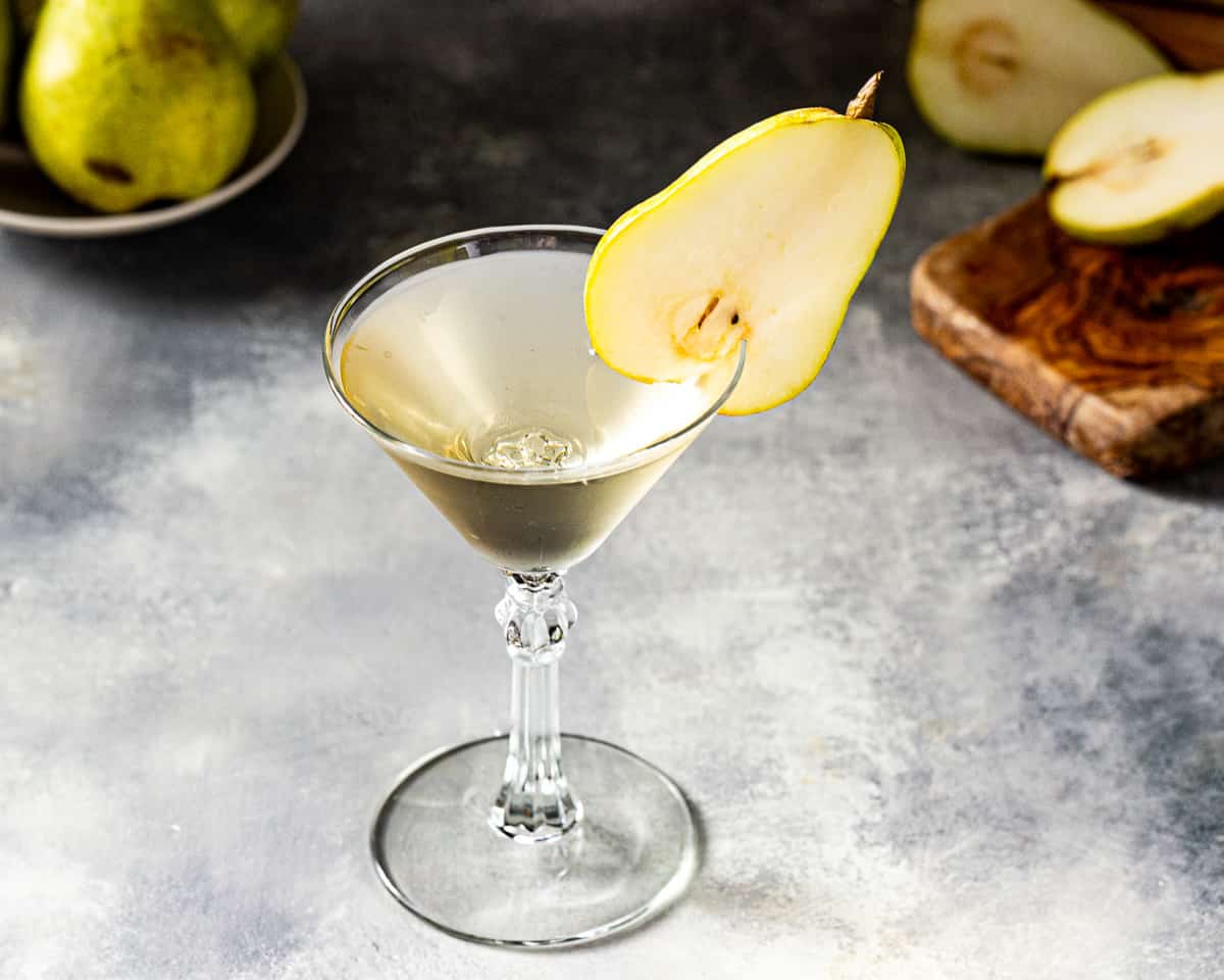 Overhead view of a Pear Martini in a stemmed glass on a gray countertop. In the background are fresh pears and a cutting board.