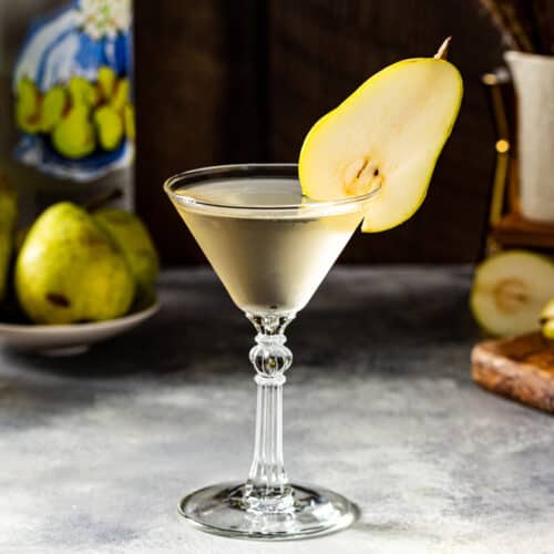 Side view of a Pear Martini cocktail in a stemmed martini glass on a gray countertop. A sliced pear is balanced on the rim of the glass as a garnish. In the background is a bottle of pear vodka along with fresh pears on the left, and on the right is a flower vase, gold colored jigger and cut pears on a cutting board.