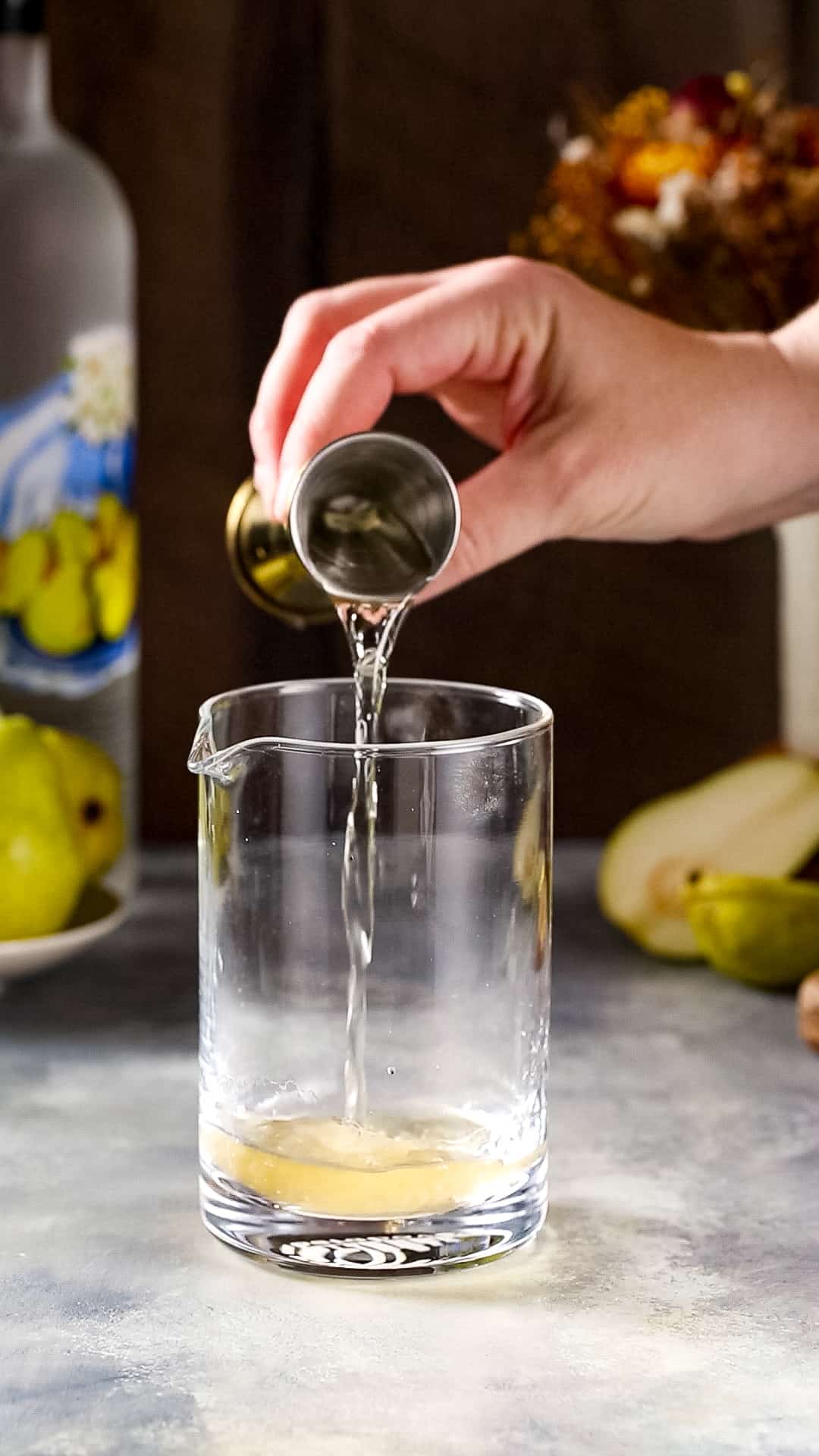 Hand pouring dry vermouth into a cocktail mixing glass from a gold jigger.