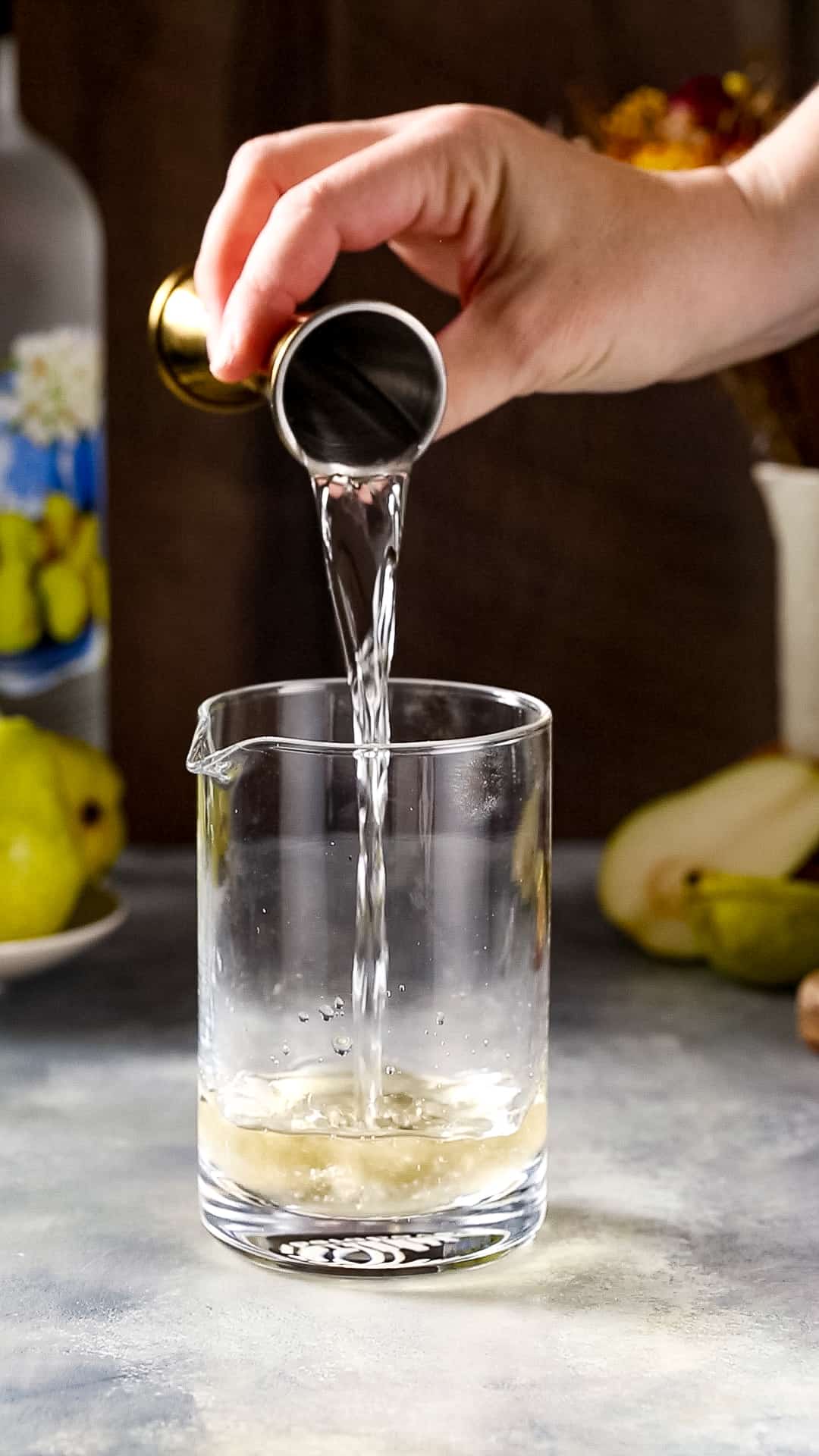 Hand pouring pear flavored vodka into a cocktail mixing glass from a gold jigger.