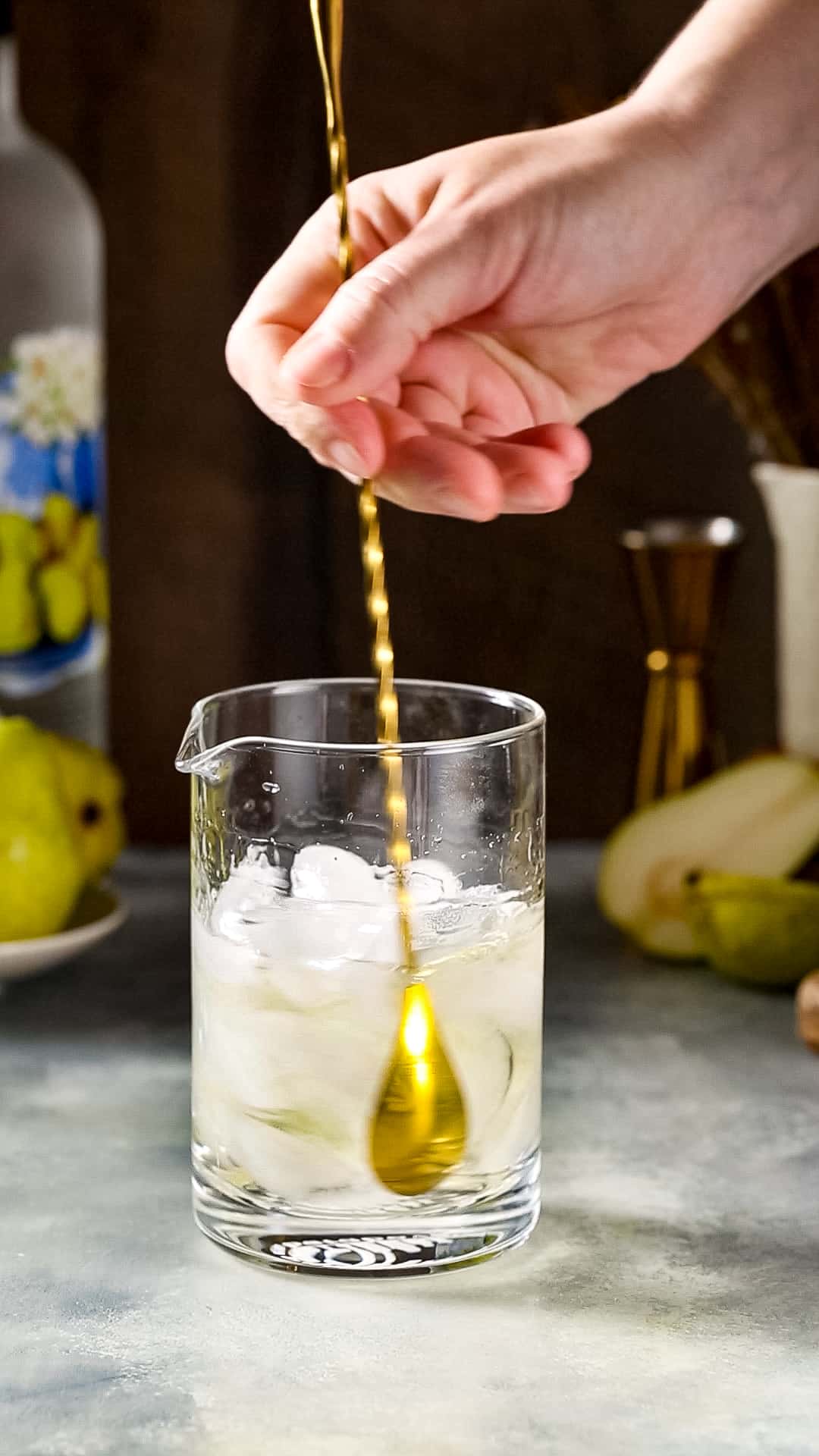 Hand holding a gold bar spoon that is stirring a cocktail along with ice in a mixing glass.