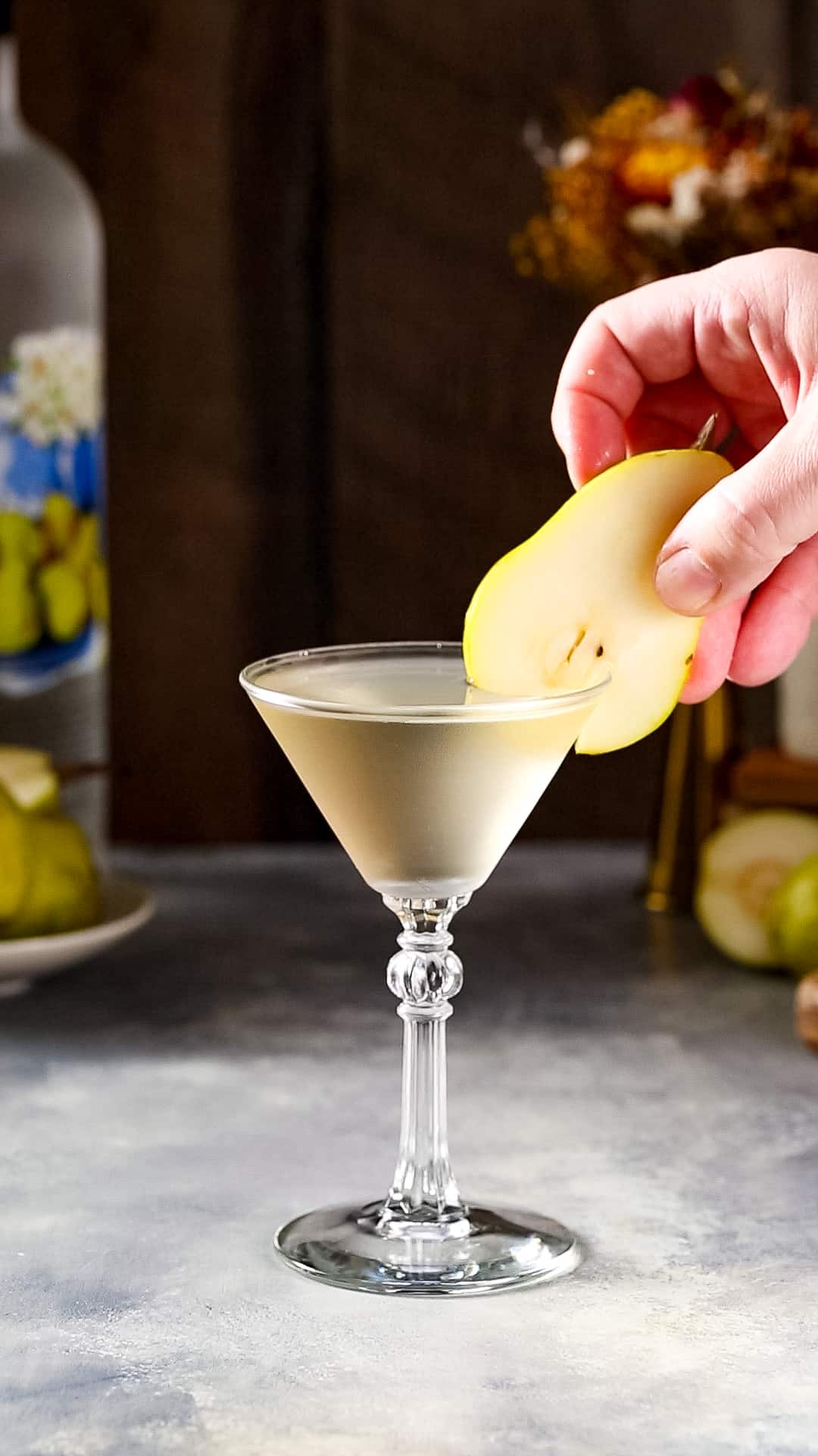 Hand adding a fresh pear slice to the rim of a martini glass filled with a light yellow liquid as a garnish.