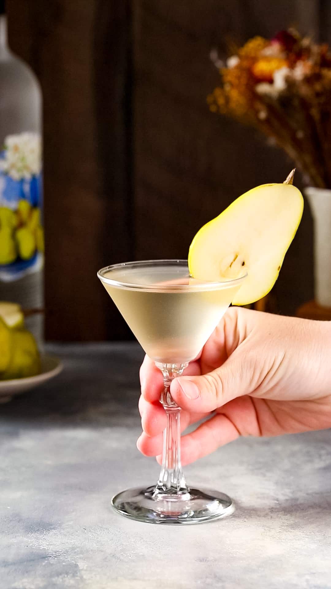Hand about to pick up a martini glass filled with a light yellow liquid that has a fresh pear slice as a garnish.