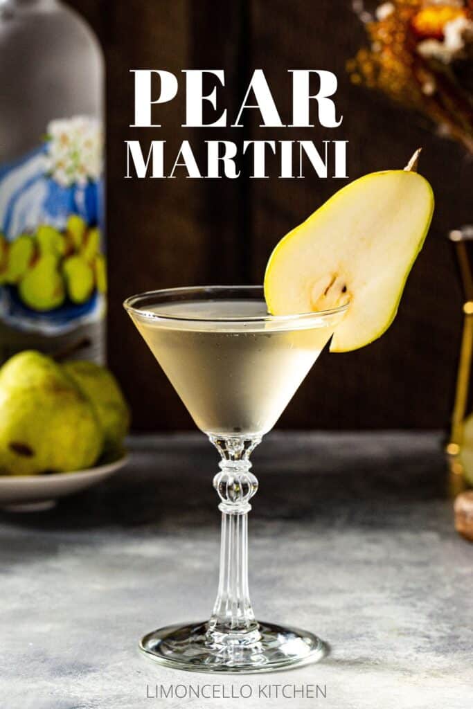 Side view of a Pear Martini cocktail in a stemmed martini glass on a gray countertop. A sliced pear is balanced on the rim of the glass as a garnish. In the background is a bottle of pear vodka along with fresh pears on the left, and on the right is a flower vase, gold colored jigger and cut pears on a cutting board. Text above the drink reads “Pear Martini”.