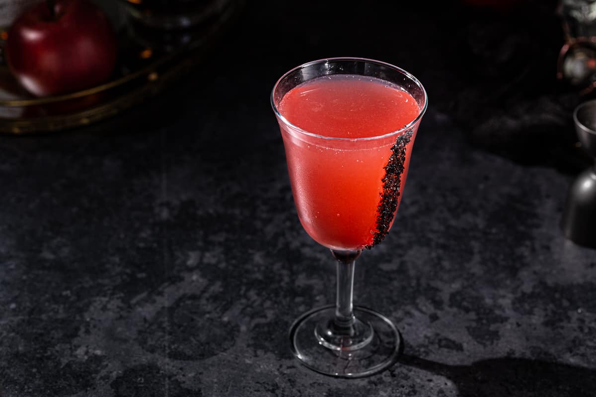 Overhead view of a red Vampire's kiss cocktail with black lava salt stuck to the side of the glass. The glass is on a dark tabletop and there are some cocktail tools around it, although they are dark and the brightest thing is the red drink.