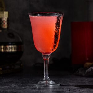 Side view of a Vampire's Kiss cocktail in a goblet glass. The drink is red-colored and there is black lava salt on the side of the glass. The background is dark, but a bottle of Chambord and a red candle are visible.