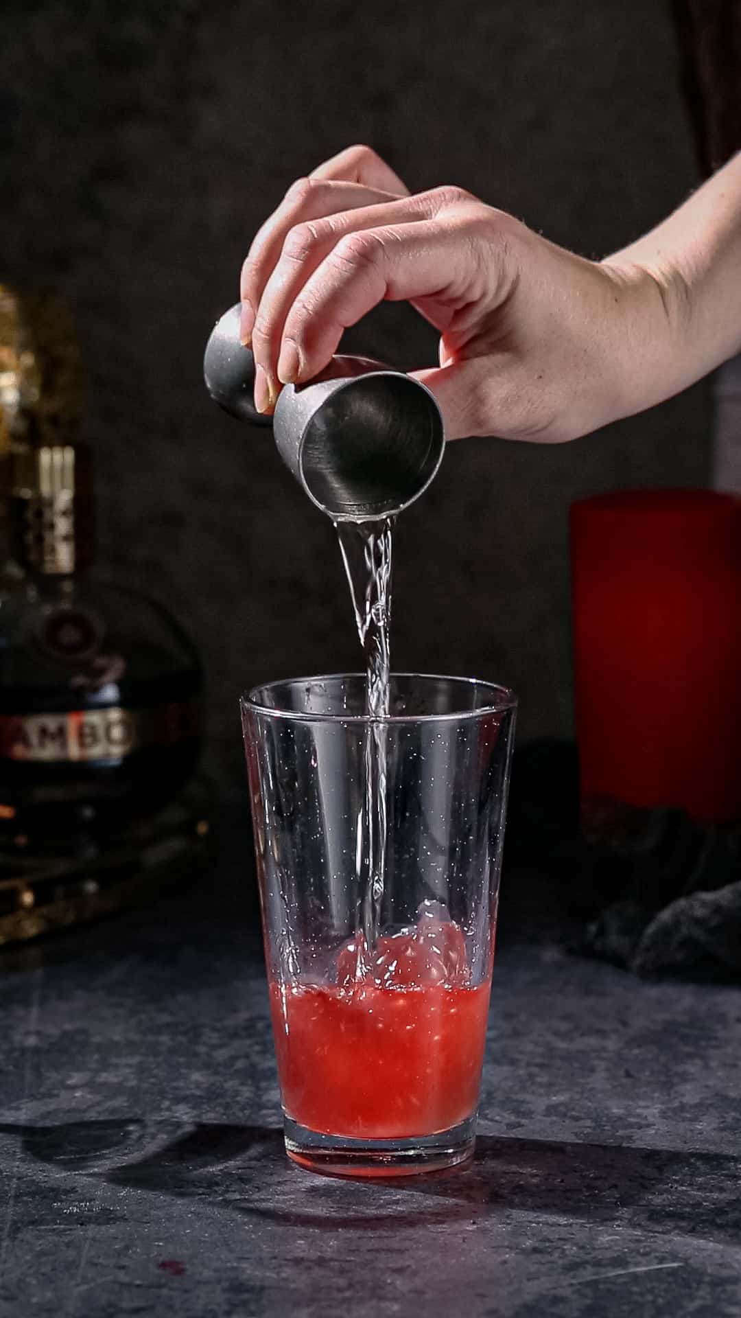 Hand adding vodka from a jigger into a cocktail shaker filled with red liquid.