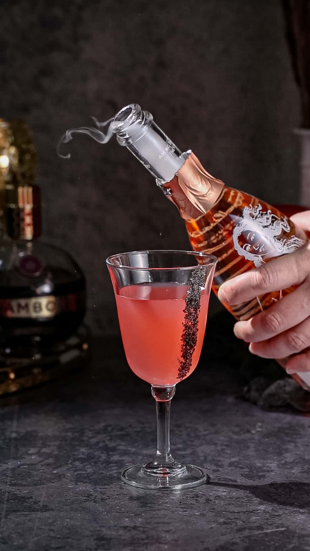 Hand holding a bottle of sparkling rose wine with some smoke coming out of it after it was freshly opened. A red cocktail in a goblet glass is in front of the bottle.