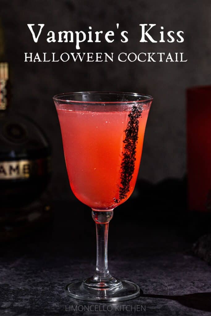 Side view of a Vampire's Kiss cocktail in a goblet glass. The drink is red-colored and there is black lava salt on the side of the glass. The background is dark, but a bottle of Chambord and a red candle are visible. Text above the drink reads "Vampire's Kiss Halloween Cocktail".