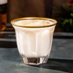 A Brandy Milk Punch cocktail with ice and freshly grated nutmeg in a faceted gold-rimmed Old Fashioned glass on a dark green countertop. In the background, part of a bottle of Armagnac is visible on the left, and on the right are some evergreen branches.