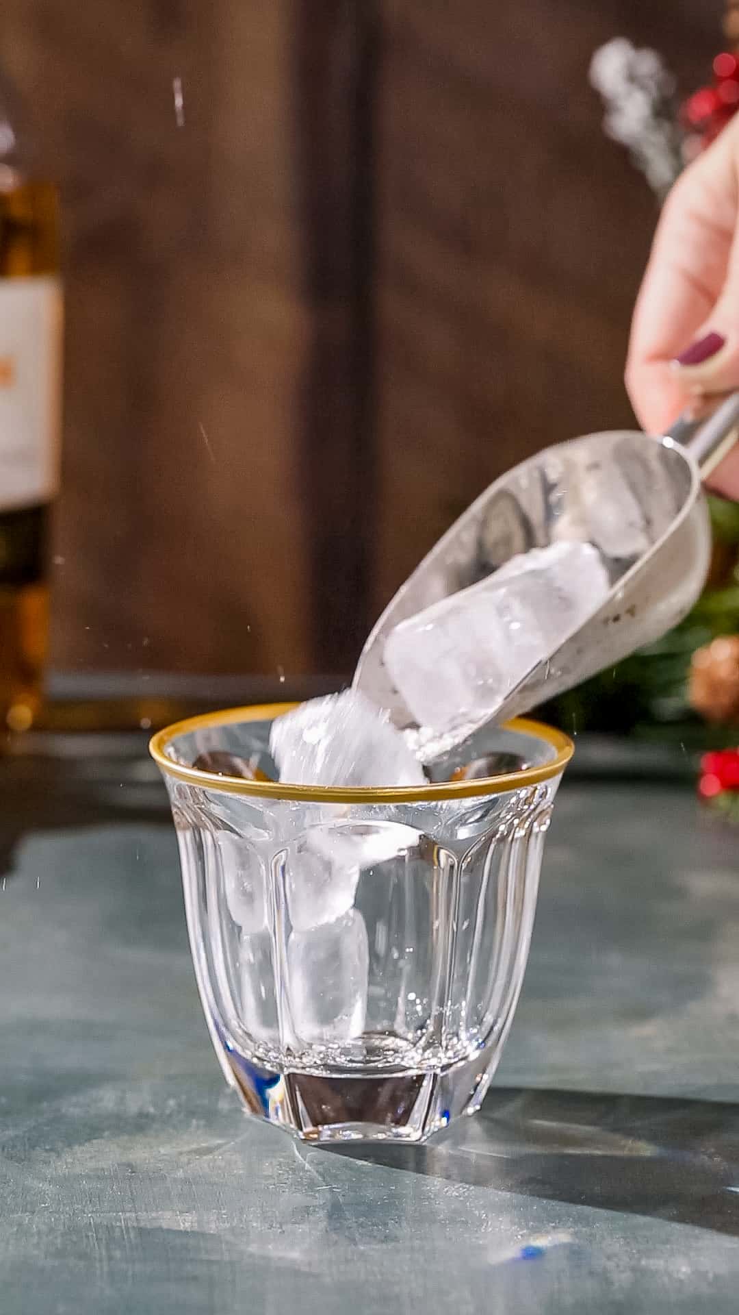 Hand using an ice scoop to add ice to a gold-rimmed old fashioned glass.