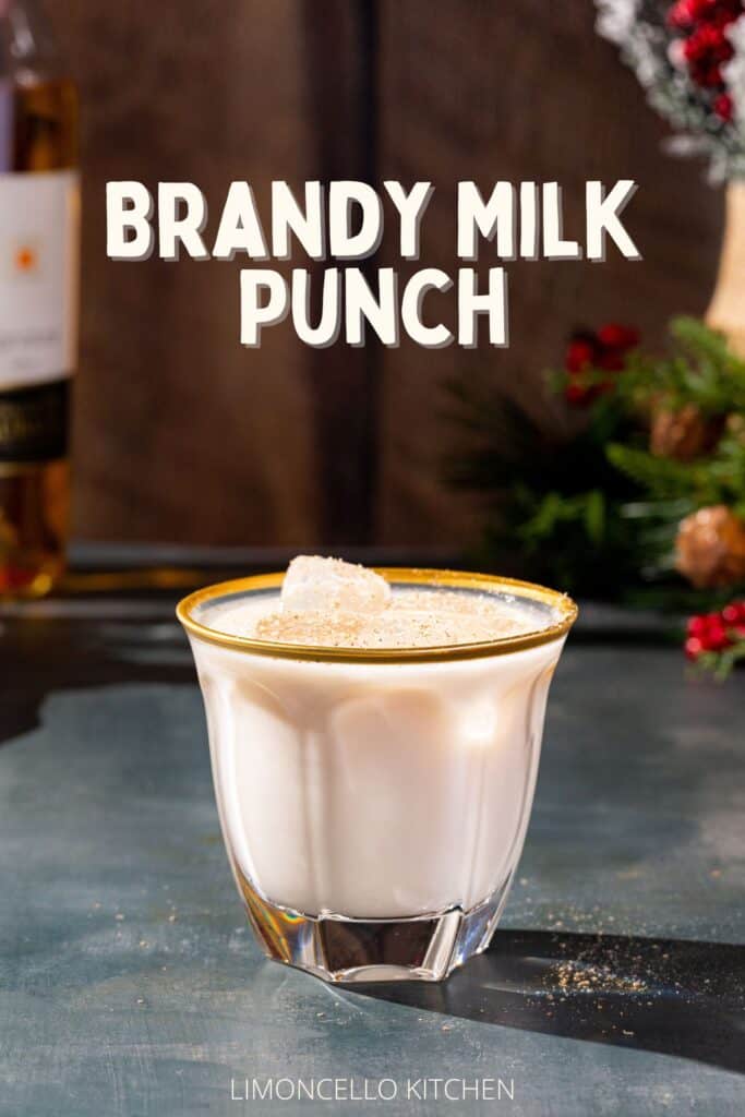 A Brandy Milk Punch in a faceted gold-rimmed Old Fashioned glass on a dark countertop. In the background, a bottle of Armagnac is visible on the left, and on the right are some evergreen branches, pinecones and winter berries. A text overlay towards the top of the image reads “Brandy Milk Punch” in the same cream color of the drink.