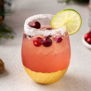 Square image of a Cranberry Margarita with a white salt rim, a lime wheel garnish and cranberries floating on top. Fresh cranberries and evergreen decor are in the background.