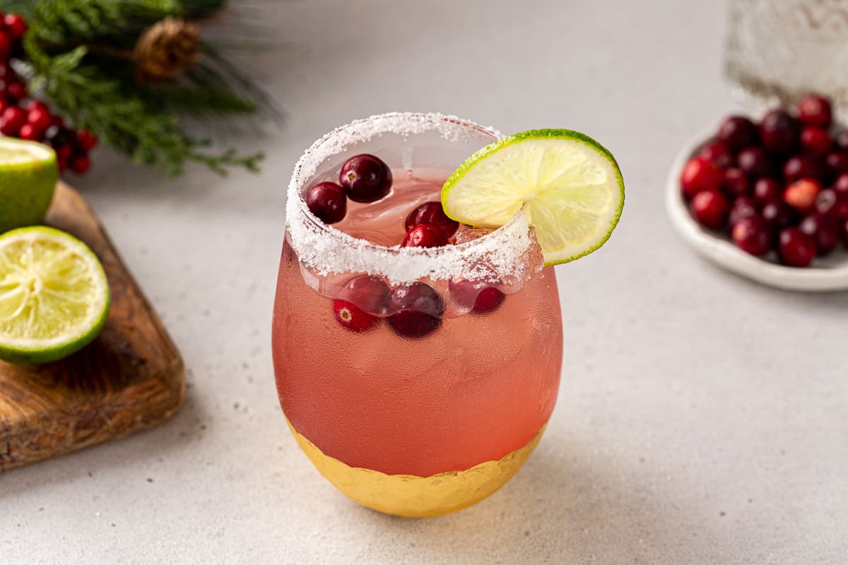 Slightly overhead shot of a Cranberry Margarita in a gold-bottomed glass with cut limes on a cutting board, pine bough decor and a plate of cranberries next to it.