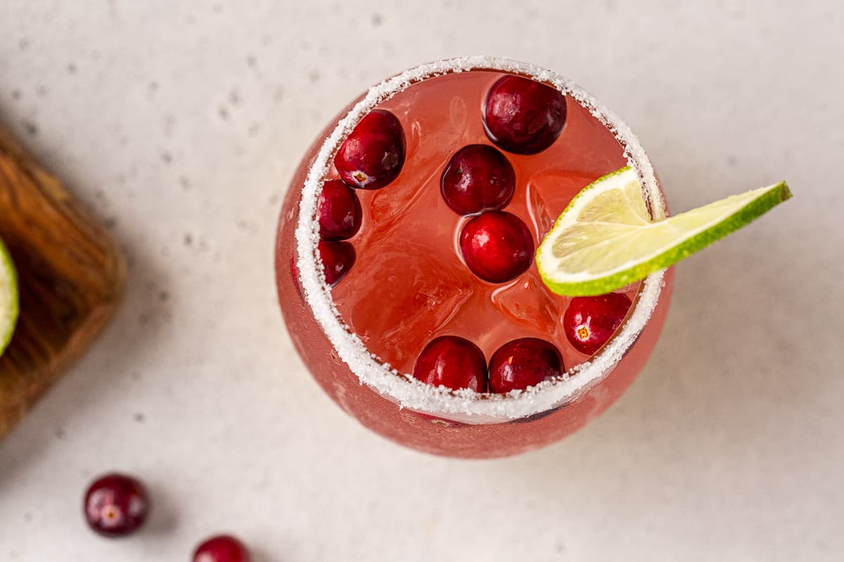 Overhead view looking down into a salt-rimmed cocktail glass filled with a cranberry margarita. A lime wheel is a garnish on the rim of the glass and fresh cranberries are floating on top.