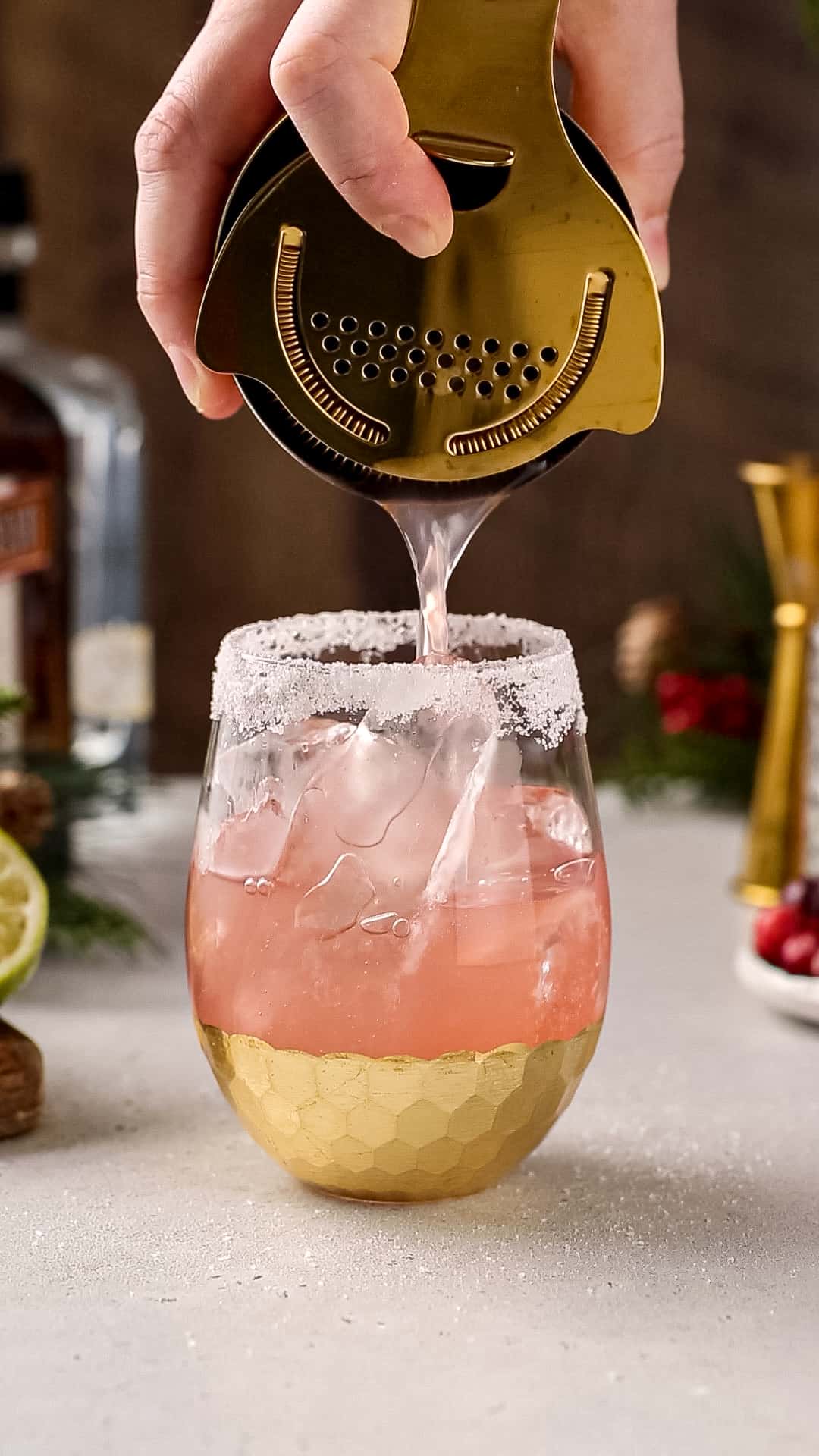 Hand straining a red-colored cocktail into an ice-filled, salt-rimmed glass.