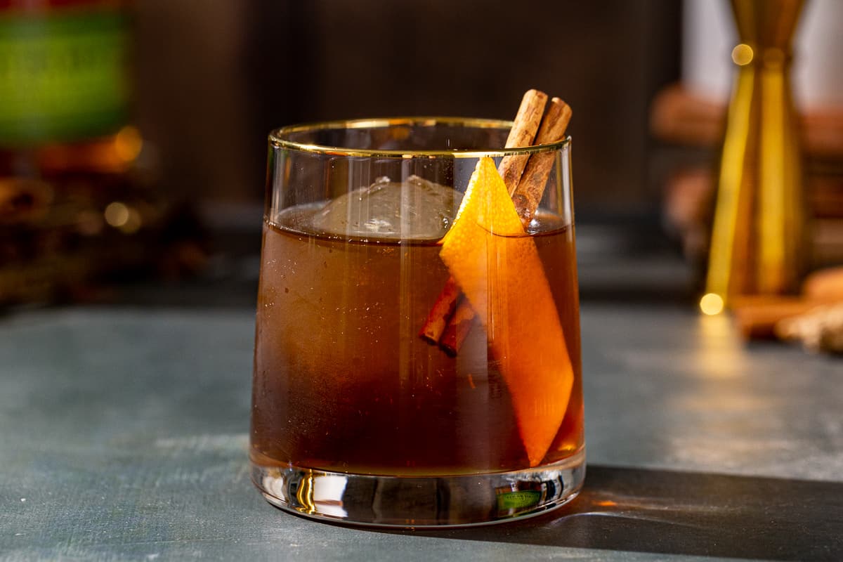 Side view closeup of a Gingerbread Old Fashioned cocktail in an old fashioned style glass, also called a lowball glass. The drink is brown and has a large sphere of ice in the glass along with an orange peel and a cinnamon stick garnish. In the background are a bottle of Bulleit rye whiskey and a gold jigger.