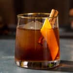 Side view of a Gingerbread Old Fashioned cocktail in an old fashioned style glass, also called a lowball glass. The drink is brown and has a large sphere of ice in the glass along with an orange peel and a cinnamon stick garnish.