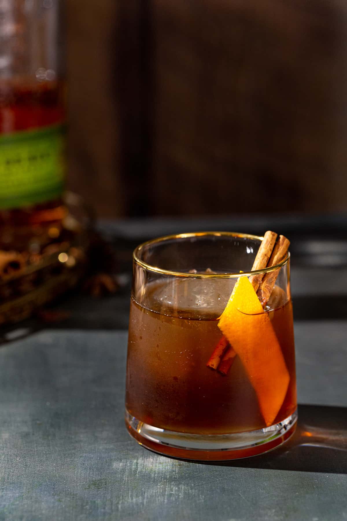 Gingerbread Old Fashioned cocktail in an old fashioned style glass. The drink is brown and has a large sphere of ice in the glass along with an orange peel and a cinnamon stick garnish. In the background is a bottle of Bulleit rye whiskey.