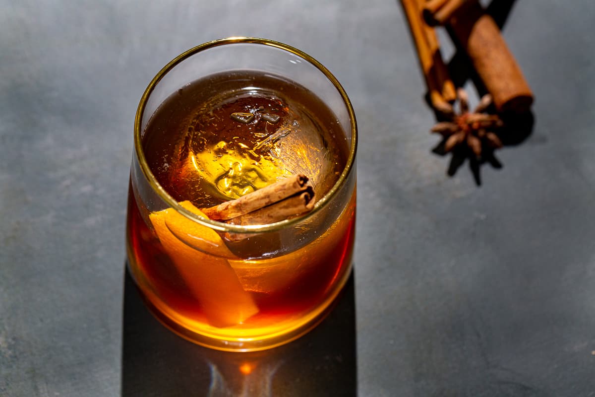 Overhead view of Gingerbread Old Fashioned cocktail in an old fashioned style glass. The drink is brown, but light coming through the drink makes it look like a range of colors from amber to dark brown. A large sphere of ice is in the glass along with an orange peel and a cinnamon stick garnish. To the right are cinnamon sticks and star anise.