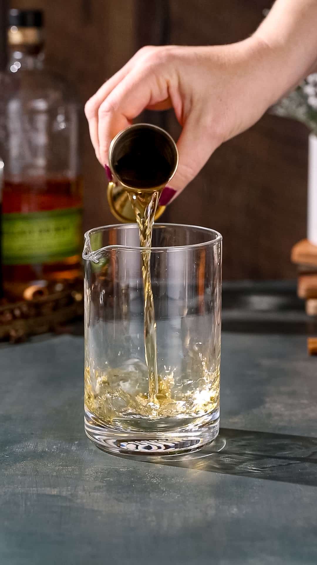 Hand pouring rye whisky into a cocktail stirring glass using a gold jigger.