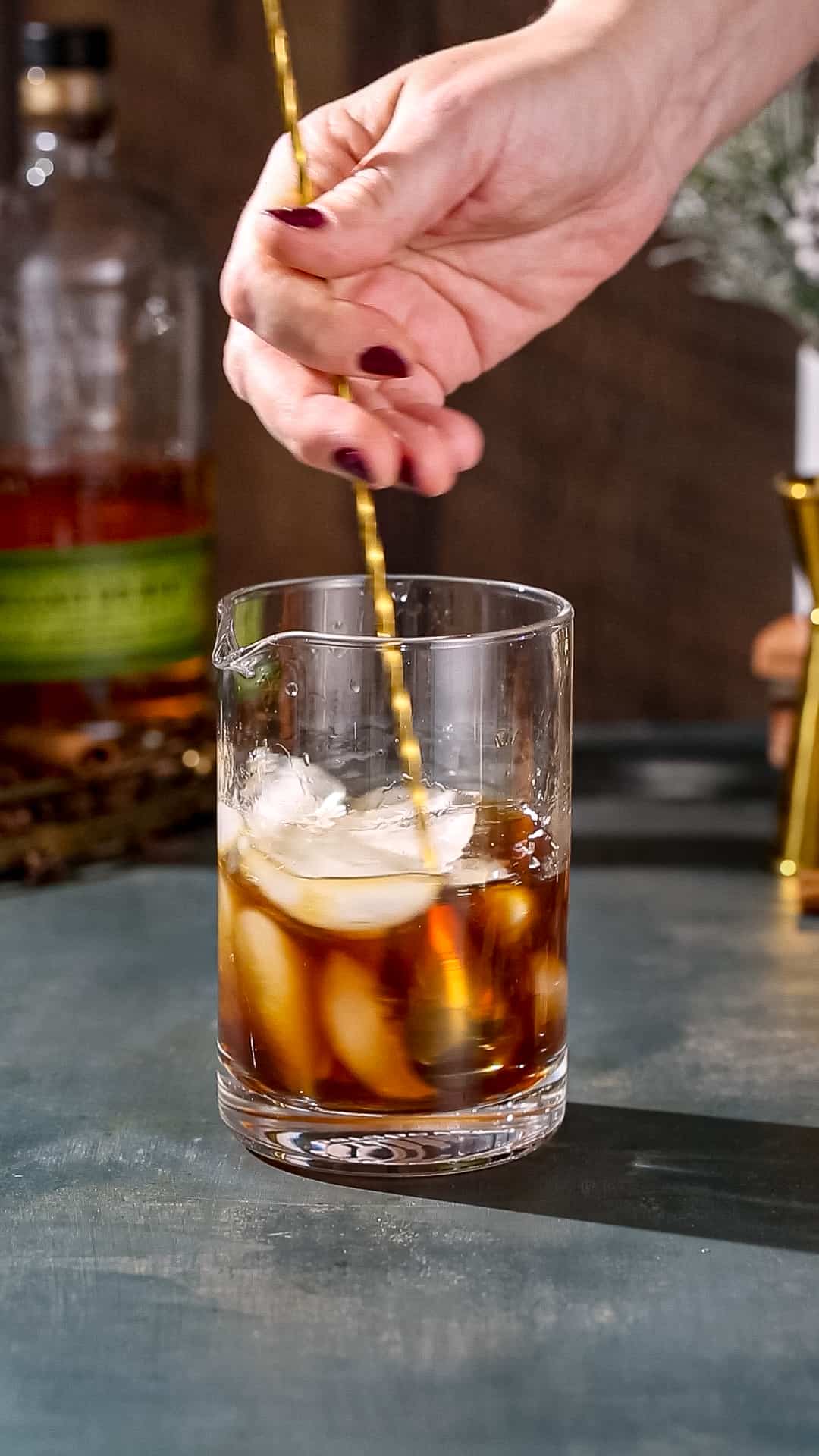 Hand using a gold bar spoon to stir a brown drink with ice in it.