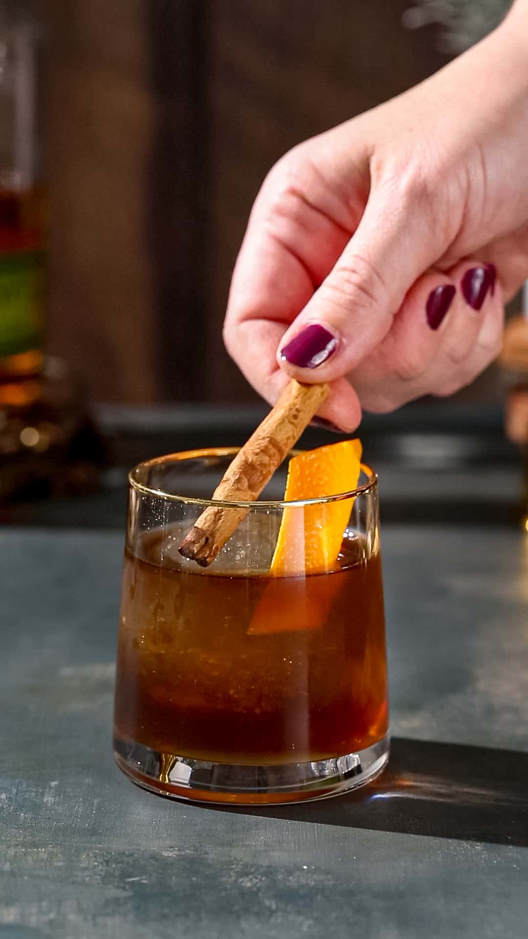 Hand adding a whole cinnamon stick to a brown cocktail that also has a large ice sphere and a parallelogram shaped orange peel in it.