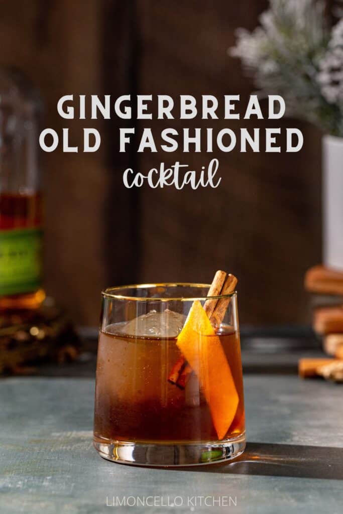 Gingerbread Old Fashioned cocktail in an old fashioned style glass. The drink is brown and has a large sphere of ice in the glass along with an orange peel and a cinnamon stick garnish. In the background to the left is a bottle of Bulleit rye whiskey, and to the right is a vase with snowy looking evergreens with a stack of wooden coasters under the vase. Text overlay above the cocktail reads “Gingerbread Old Fashioned cocktail”.