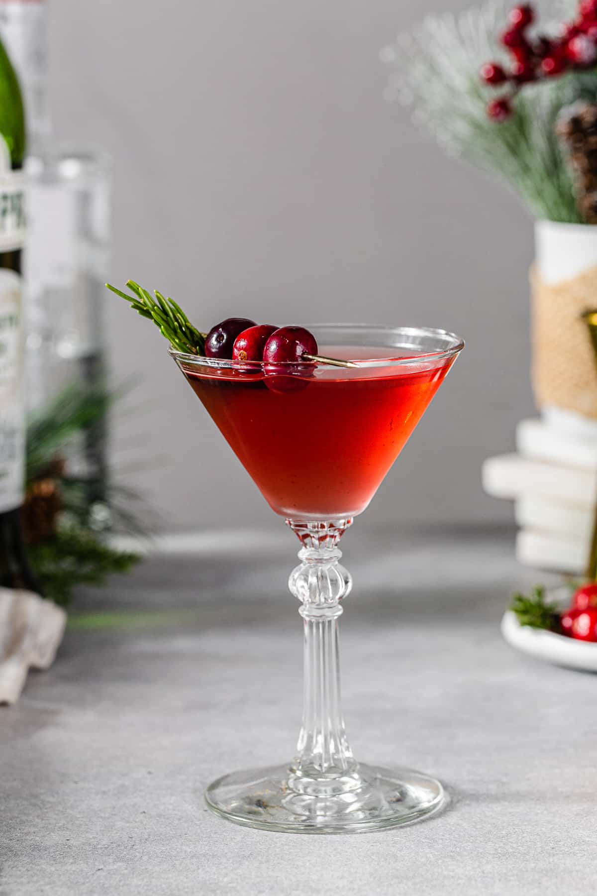 Side view of a Mistletoe Martini cocktail in a martini style glass. The red colored cocktail is garnished with three cranberries on a gold cocktail pick and a sprig of fresh rosemary. To the right of the glass is a plate of cranberries and rosemary along with a gold jigger. On the left are evergreen sprigs, a bottle of gin and a green bottle of vermouth.